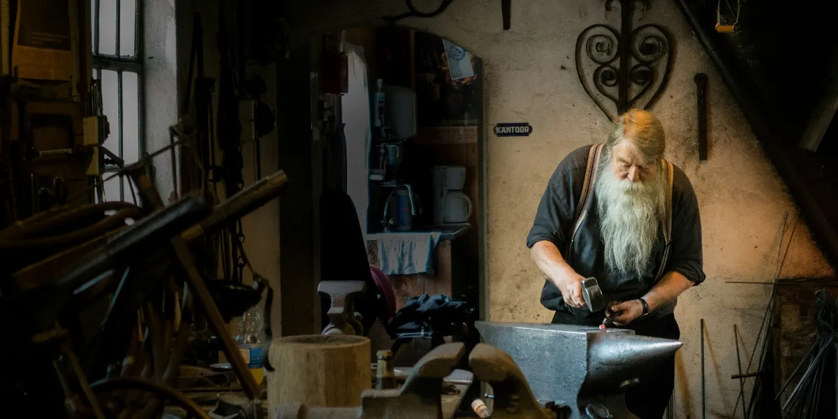 A white-bearded blacksmith hammers metal on an anvil in a dim workshop full of tools