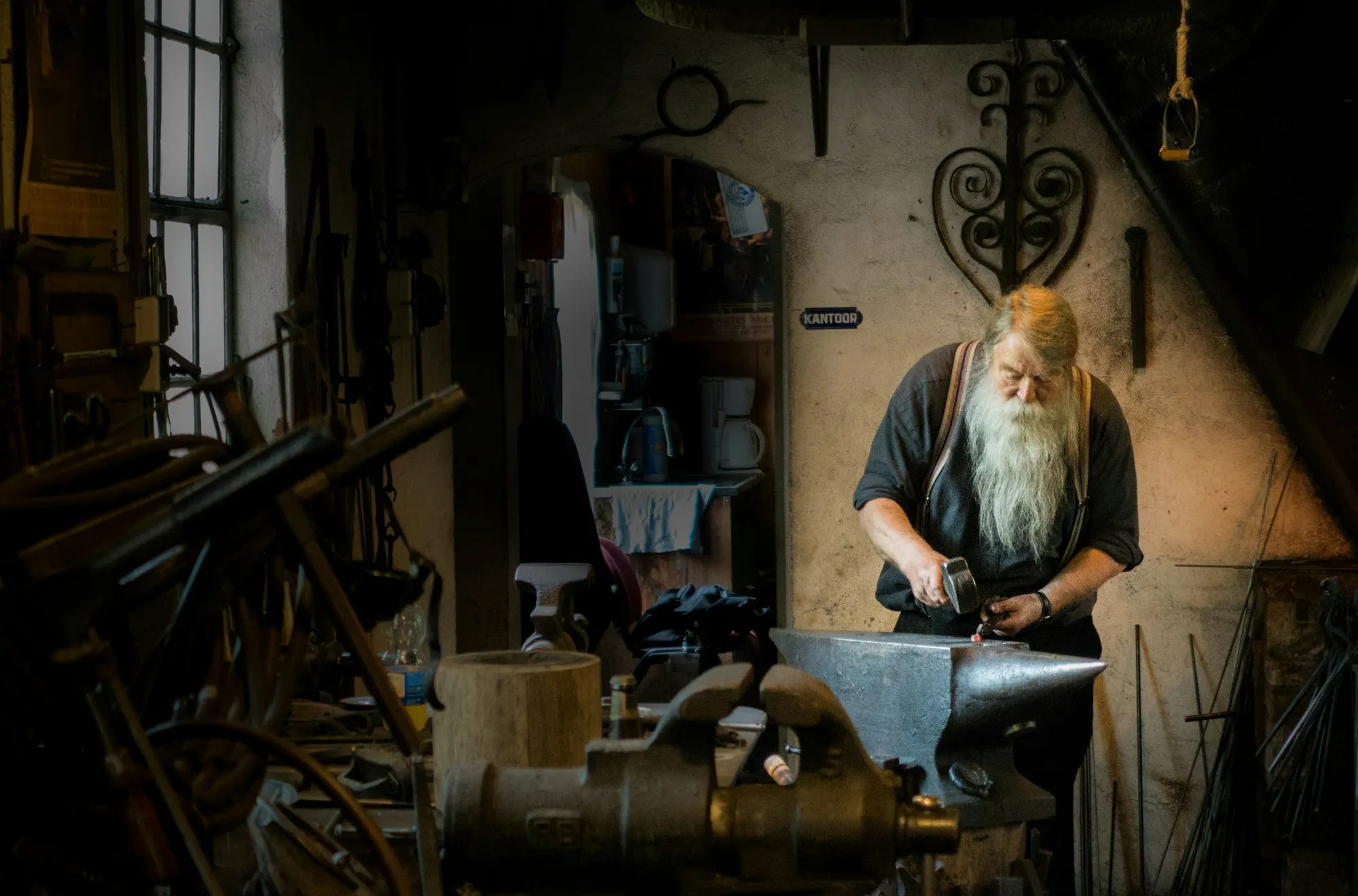 A white-bearded blacksmith hammers metal on an anvil in a dim workshop full of tools
