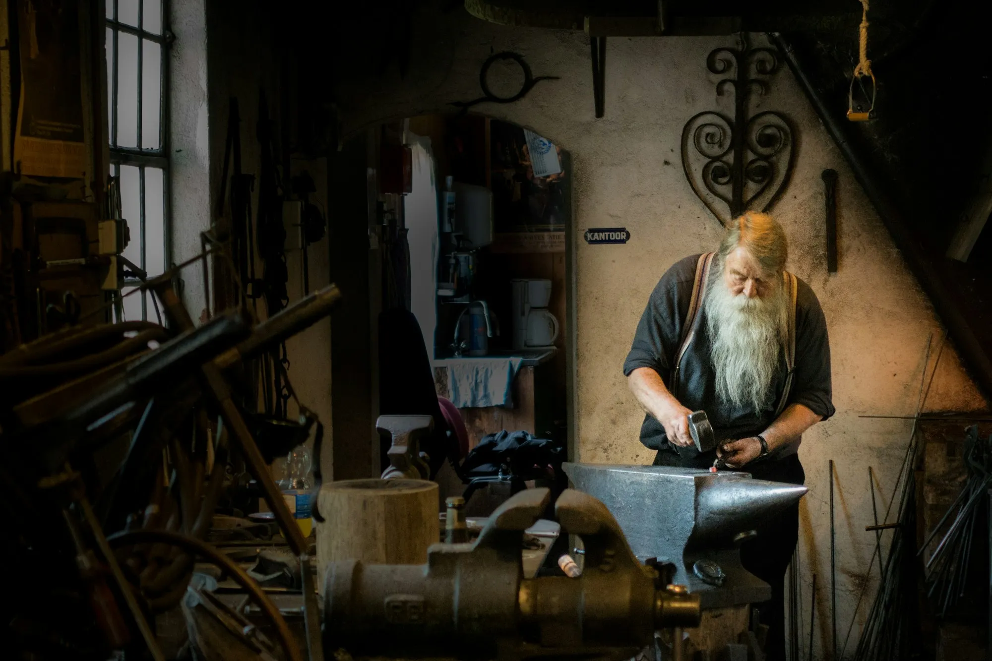 A white-bearded blacksmith hammers metal on an anvil in a dim workshop full of tools